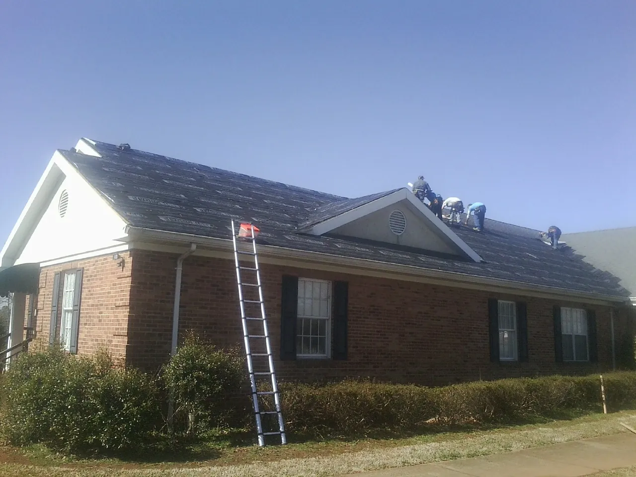 Crew tearing shingles off a brick ranch roof with a ladder set — Metal Roof Repair work by our crew in Leeds